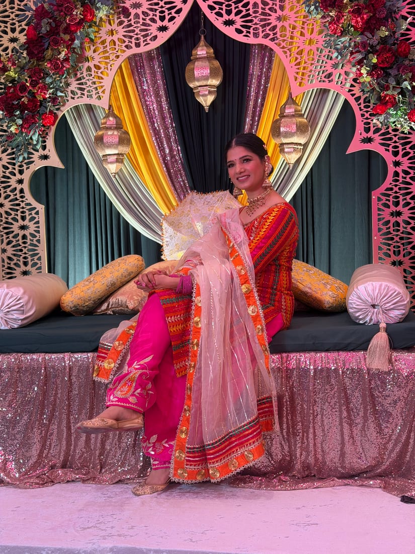 A guest in a pink and orange embroidered suit seated on a decorated stage at a Punjabi wedding celebration