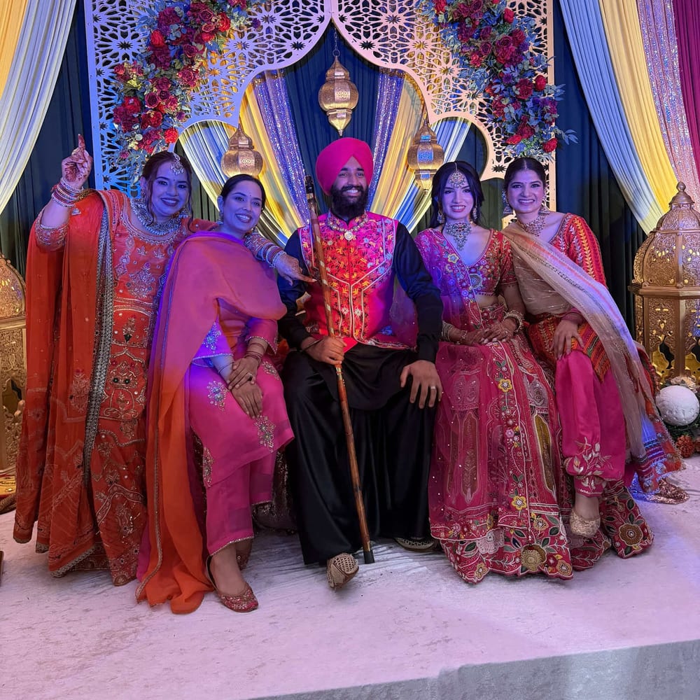 A groom in a pink turban and embroidered jacket seated with four family members in red, pink, and orange suits, on a stage framed by a floral backdrop