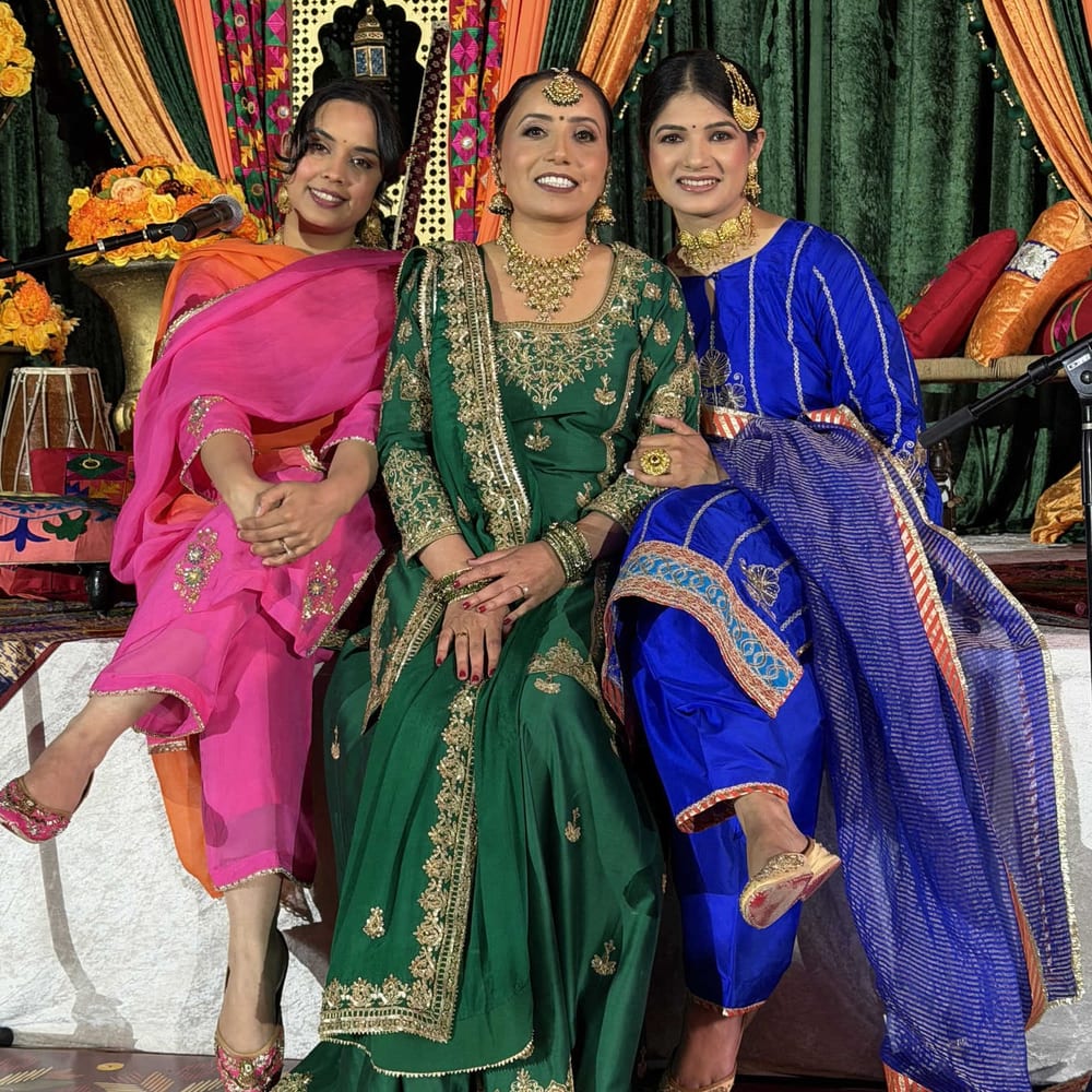 Three women in pink, green, and blue embroidered suits with gold jewellery, seated together on a decorated Sangeet stage