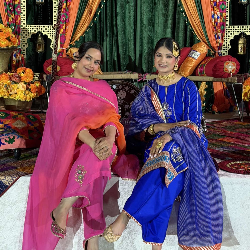 Two women in pink and royal blue embroidered suits seated on a decorated Sangeet stage, framed by orange and green drapes and marigold arrangements