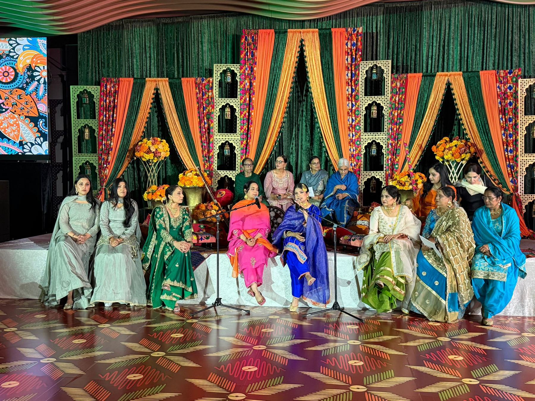Women in embroidered suits seated together on a decorated Sangeet stage, two of them singing at a microphone in front of orange and green drapes and marigold arrangements