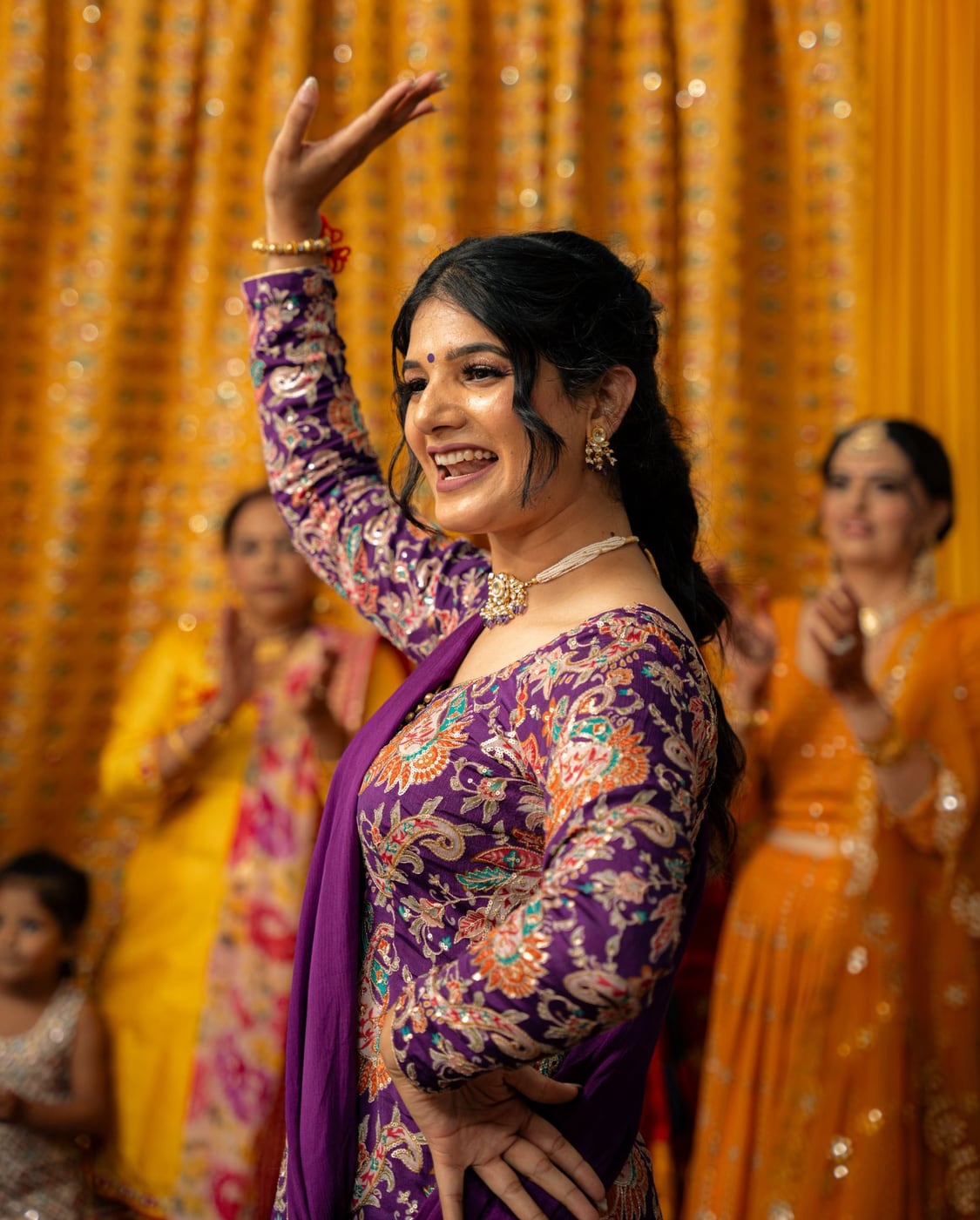Harpreet Sidhu dancing Gidha in a purple embroidered suit, one arm raised, with guests clapping behind her